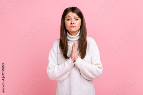 Female holds hands in prayer, looking with imploring pleading expression, begging help, forgiveness, wearing white casual style sweater. Indoor studio shot isolated on pink background.
