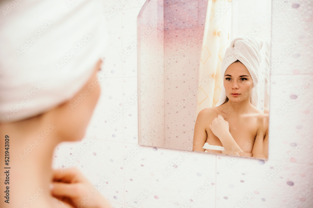 Portrait of young girl with towel on head in white bathroom looks and touches her face in the mirror and enjoys youth and hydration. Natural beauty, home care for problem skin Portrait of young girl with towel on head in white bathroom looks and touches her face in the mirror and enjoys youth and hydration. Natural beauty, home care for problem skin