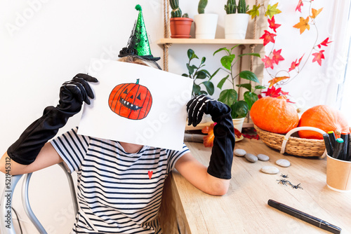A child shows his drawing for Halloween, sitting at a desk by the window, surrounded by pumpkins and houseplants. Sketch drawing of an orange laughing pumpkin with sharp teeth. Get ready for the party