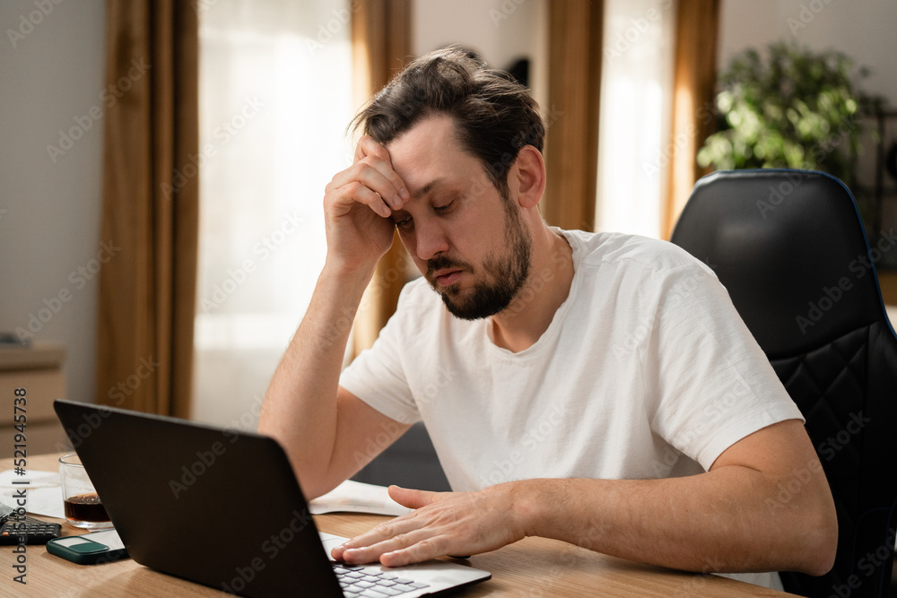 A tired middle-aged man works at his workplace. He rested head on his arm. The man looked thoughtfully at the laptop screen and stopped with hand on the keyboard. On the table is phone, calculator. A tired middle-aged man works at his workplace. He rested head on his arm. The man looked thoughtfully at the laptop screen and stopped with hand on the keyboard. On the table is phone, calculator.