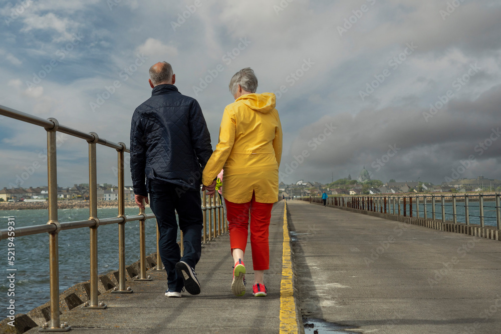 Elderly couple walking by the ocean holding hands, back to viewer. Man in dark clothes, women in bright saturated yellow jacket an red trousers. Cloudy sky. Town in the background. Romantic scene.