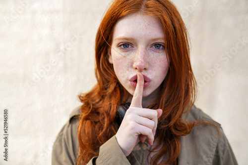 Hipster teen gen z redhead girl showing shh sign finger gesture asking to keep secret, be hush silent or privacy silence on urban wall background. Teenage problem secrecy concept. Close up portrait photo