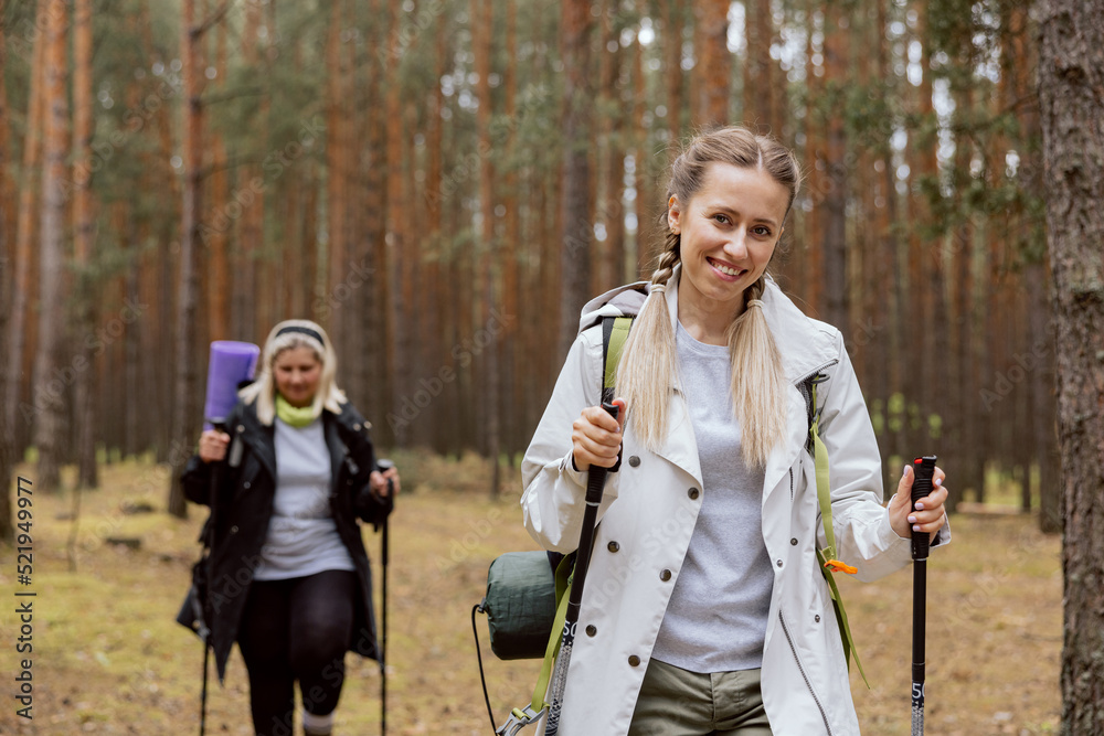 Mother and daughter walking in the forest. Young girl is walking ahead, elderly woman behind. Women are holding rucksack, trecking poles. Woman is smiling. Mother and daughter walking in the forest. Young girl is walking ahead, elderly woman behind. Women are holding rucksack, trecking poles. Woman is smiling.