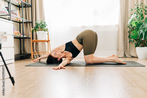 Fit woman working out, doing stretching exercise on yoga mat while making video translation fitness video online on smartphone device on tripod at home. Female practicing yoga exercise online