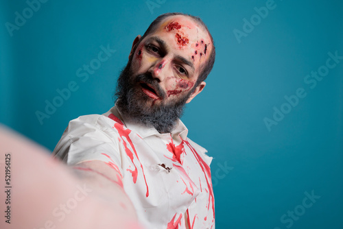 Undead zombie holding camera to take photo, looking creepy and dangerous over blue background. Scary brain eating devil with horror look and aggressive bloody face taking frightening picture.