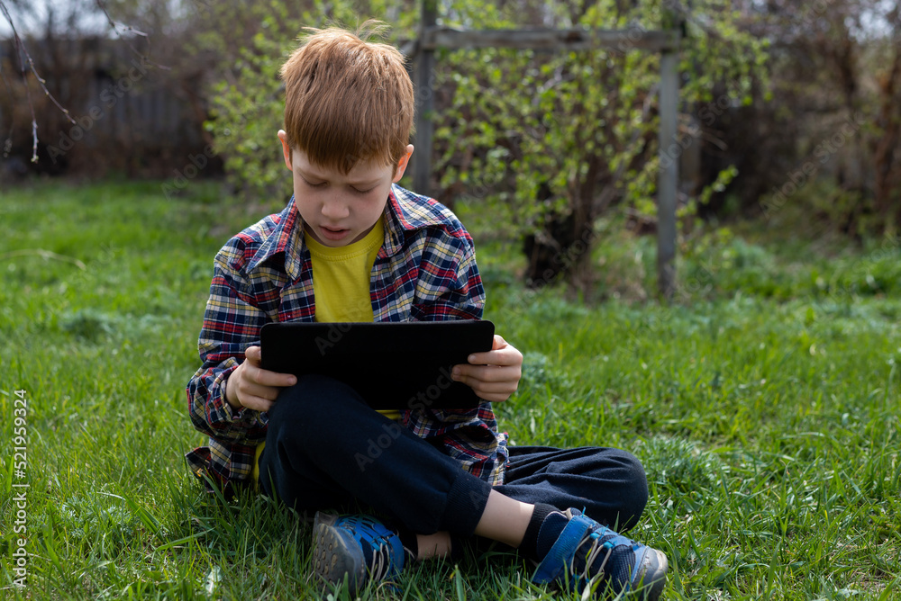 Happy child ginger boy playing on tablet or watching cartoons sitting on green grass in the backyard in the village, smiling Happy child ginger boy playing on tablet or watching cartoons sitting on green grass in the backyard in the village, smiling