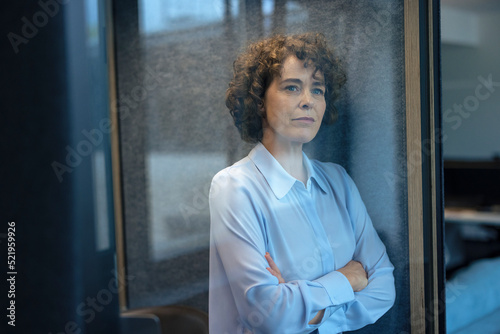 Thoughtful businesswoman standing with arms crossed seen through glass