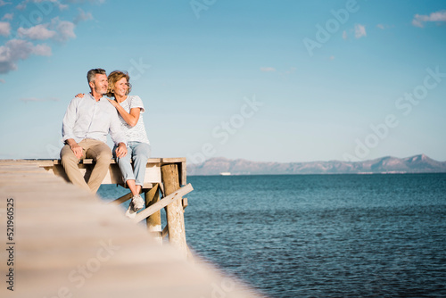 Happy mature couple sitting on jetty over sea in front of sky photo