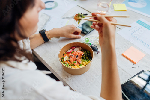 Businesswoman eating healthy poke bowl with chopsticks in lunch break at office photo