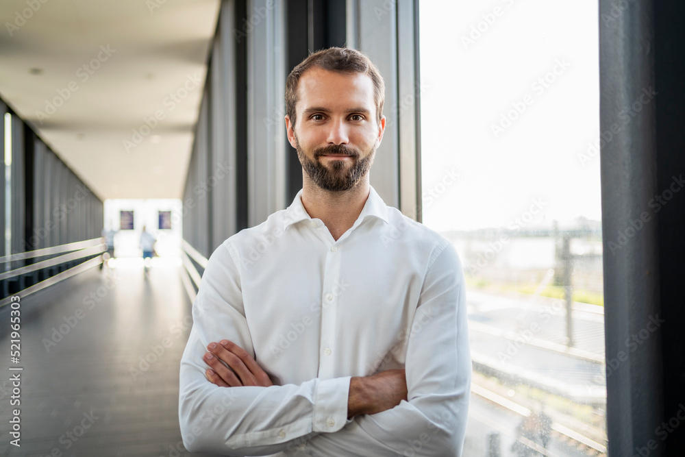 Confident young businessman with arms crossed standing by window on elevated walkway