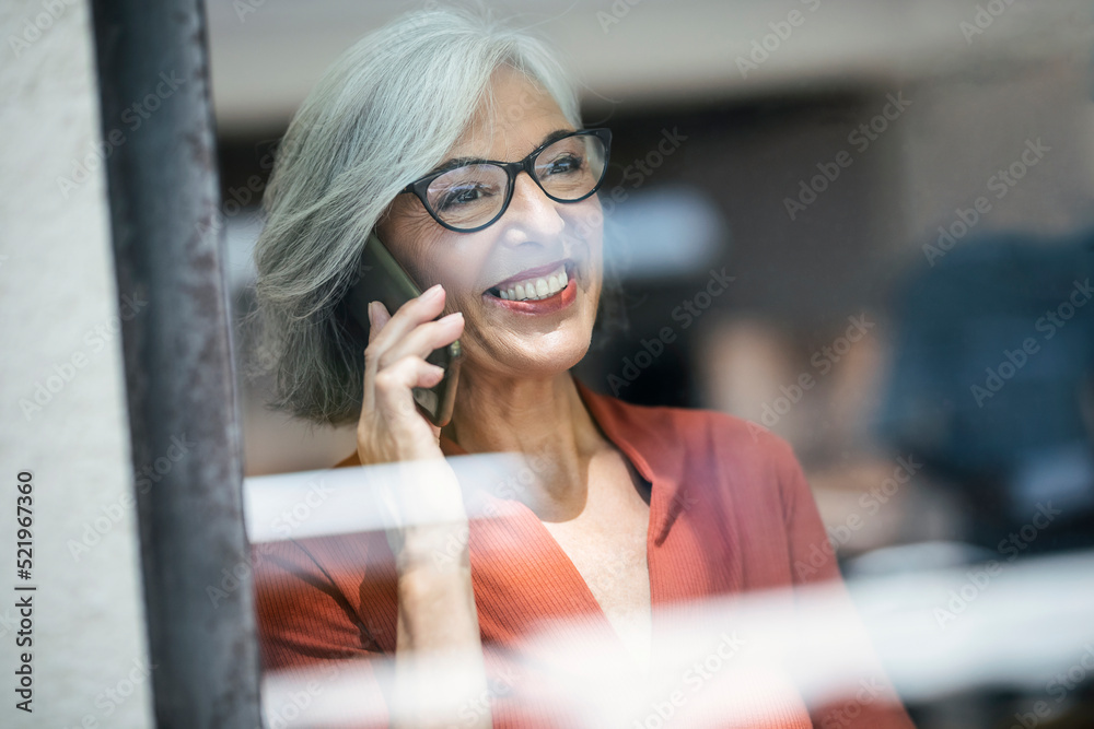 Smiling senior businesswoman talking on phone seen through glass window Smiling senior businesswoman talking on phone seen through glass window