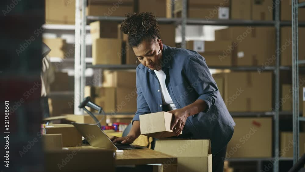 Inventory Manager Preparing a Small Cardboard Parcel for Postage. Multiethnic African American Female Small Business Owner Working on Laptop Computer in Warehouse. Inventory Manager Preparing a Small Cardboard Parcel for Postage. Multiethnic African American Female Small Business Owner Working on Laptop Computer in Warehouse.