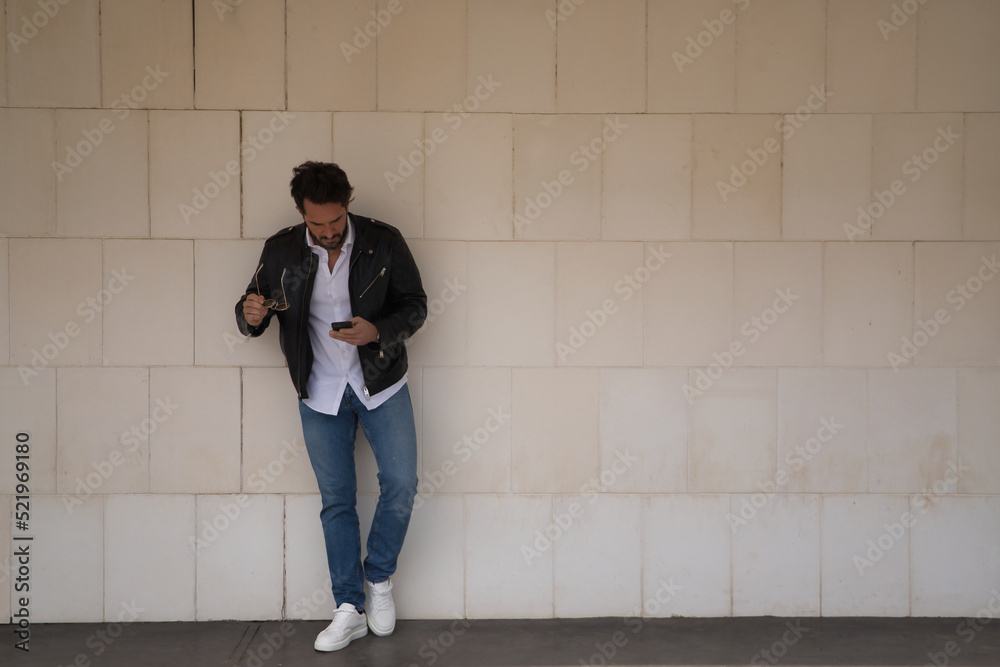 Handsome young man with beard, sunglasses, leather jacket, white shirt and jeans, leaning against a white wall, consulting his cell phone. Concept beauty, trend, smartphone, app, social networks. Handsome young man with beard, sunglasses, leather jacket, white shirt and jeans, leaning against a white wall, consulting his cell phone. Concept beauty, trend, smartphone, app, social networks.