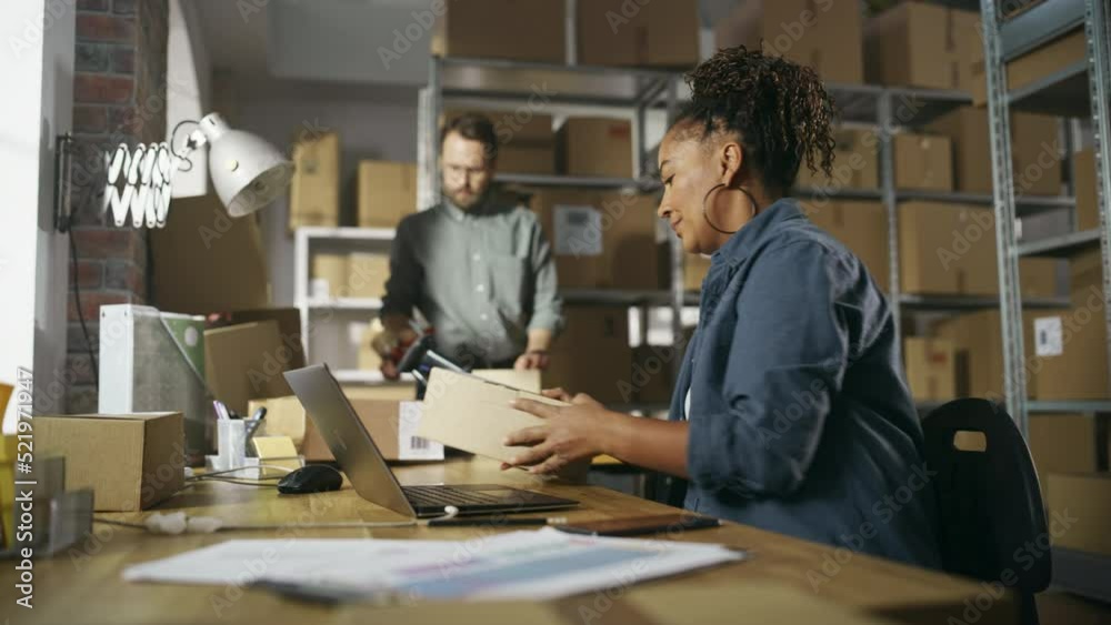 Inventory Manager Using Laptop Computer to Check Order Number on a Parcel, Preparing a Small Cardboard Box for Postage. Shelves Full of Box Packages and Two Colleagues in the Background. Inventory Manager Using Laptop Computer to Check Order Number on a Parcel, Preparing a Small Cardboard Box for Postage. Shelves Full of Box Packages and Two Colleagues in the Background.