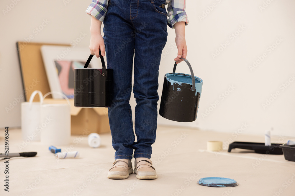 Fototapeta premium The child dressed in jeans and checked shirt holds cans of paint in hands. The girl's face is not visible, only new ones. In background are paintings, paint rollers, paint and other things for repair.