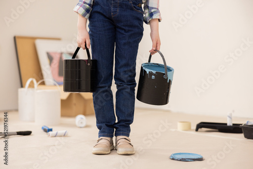 The child dressed in jeans and checked shirt holds cans of paint in hands. The girl s face is not visible  only new ones. In background are paintings  paint rollers  paint and other things for repair.