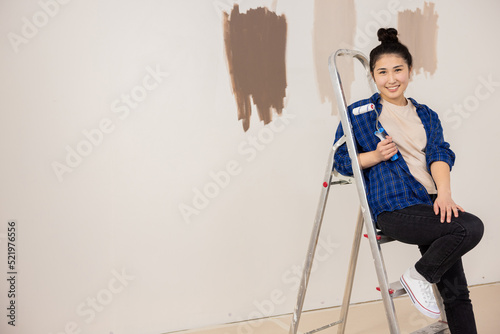 Asian woman sitting on a ladder and smiling, holding a paint roller in her hand. In the background, a wall is painted in three shades of brown. The girl is still choosing which room to paint.