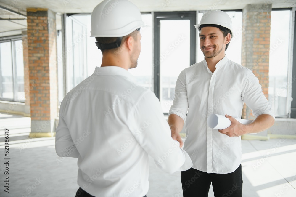 A front view of two smart architects with white helmets reviewing blueprints at a construction site on a bright sunny day A front view of two smart architects with white helmets reviewing blueprints at a construction site on a bright sunny day