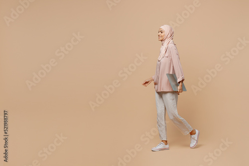 Full body side view happy young arabian asian muslim woman she wear abaya hijab pink clothes hold closed laptop pc computer isolated on plain light beige background People uae islam religious concept.
