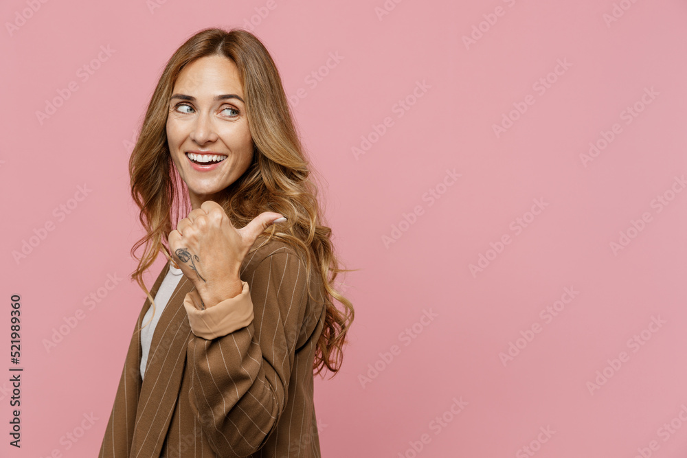 Young successful employee business woman 30s she wearing casual brown classic jacket point thumb finger aside on workspace area mock up isolated on plain pastel light pink background studio portrait. Young successful employee business woman 30s she wearing casual brown classic jacket point thumb finger aside on workspace area mock up isolated on plain pastel light pink background studio portrait.