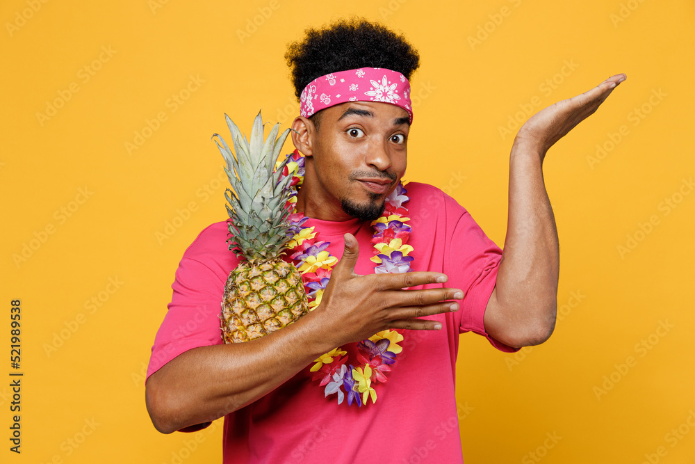 Young fun cool cheerful man 20s he wearing pink t-shirt hawaiian lei near hotel pool holding pineapple spreading hand look camera isolated on plain yellow background. Summer vacation sea rest concept.