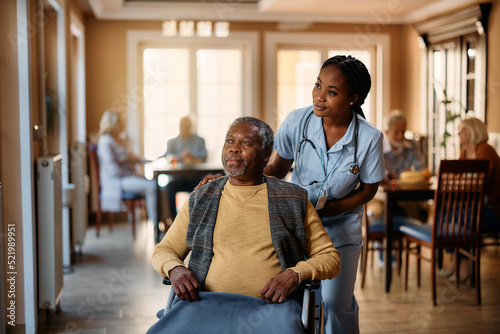 Black nurse and senior man in wheelchair look through window at nursing home.