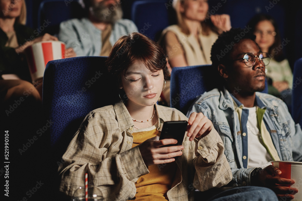 Obraz premium Young gen Z Caucasian woman getting bored with movie scrolling news feed in social network on smartphone at cinema