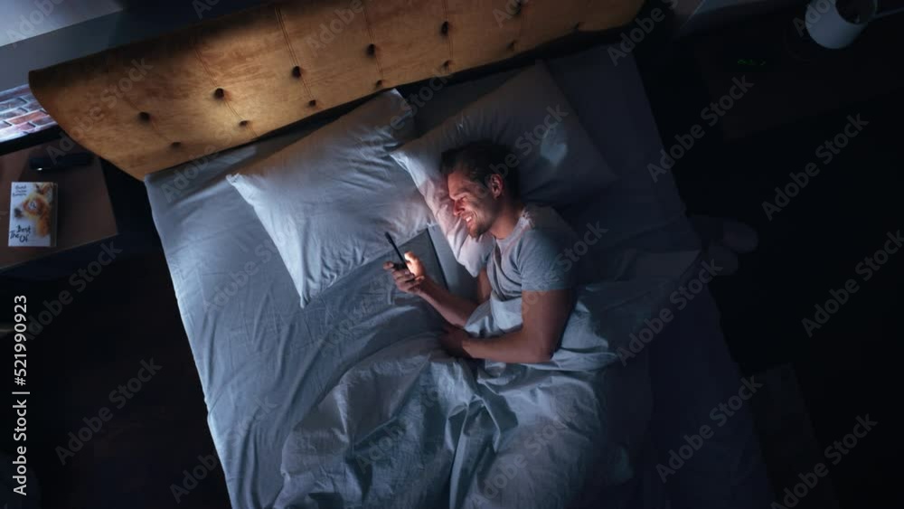 Top View Apartment Bedroom: Handsome Young Man Using Smartphone in Bed at Night. Smiling Guy Browsing Social Media, Dating Apps, Remote Work Software, Doing Internet Online Shopping on Mobile Phone Top View Apartment Bedroom: Handsome Young Man Using Smartphone in Bed at Night. Smiling Guy Browsing Social Media, Dating Apps, Remote Work Software, Doing Internet Online Shopping on Mobile Phone