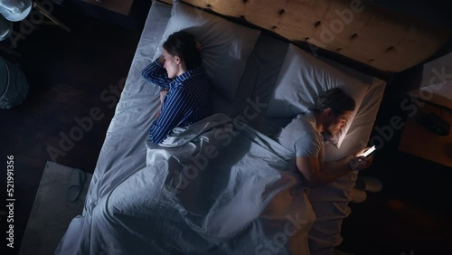 Top View Bedroom Apartment: Man Uses Smartphone in Bed at Night When His Female Partner Trying to Fall Asleep. Couple After Fight, Argument. Addictive World of Social Media, Doom Scrolling, Fake News. photo