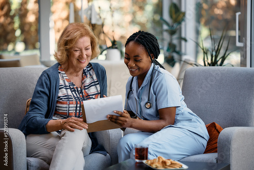 Happy senior woman and black female caregiver analyzing medical data during home visit.
