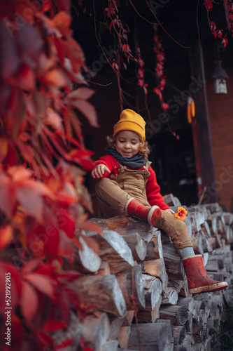 Happy little girl sitting at pile of wood with flower, surrouded by red leaves, during beautiful autumn day.