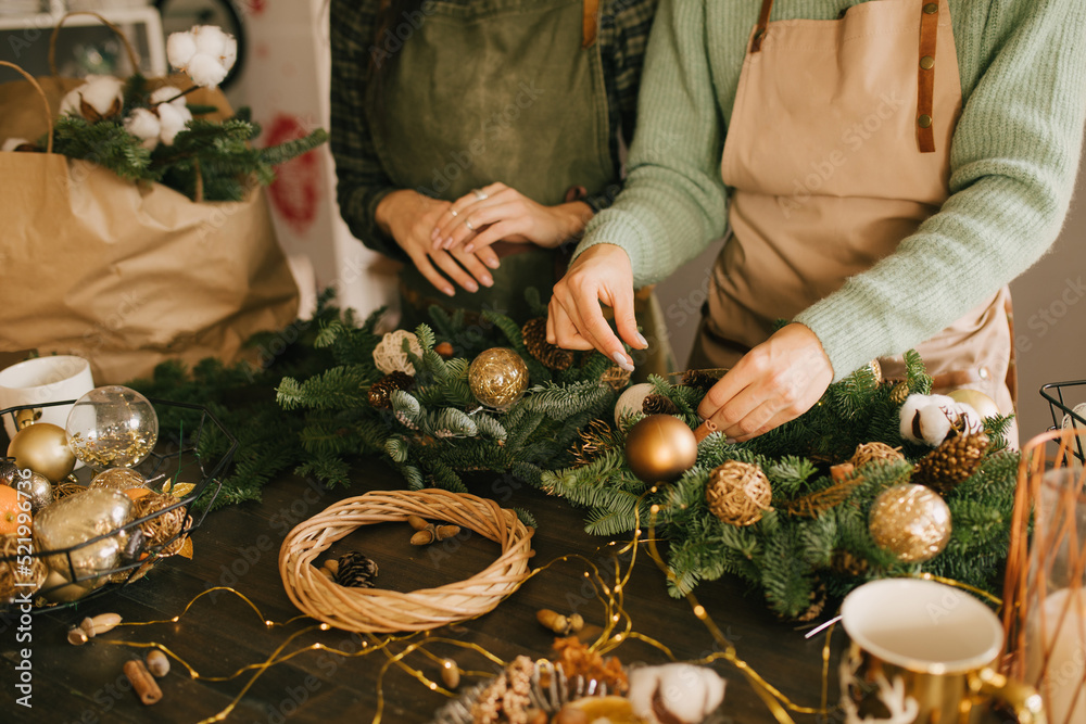 Two millennial women making Christmas wreath using pine branches and festive decorations. Small business - obrazy, fototapety, plakaty 