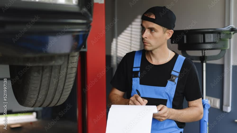 Mechanic inspects the car undercarriage way and makes a note on his inspection sheet. Automobile service, car mechanic. Car service employe inspect car. Modern workshop.