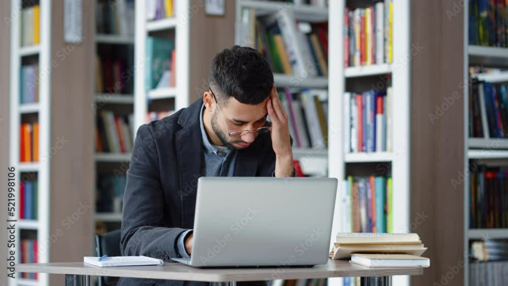 Handsome young man with eyeglasses suffering headache while working on laptop in library, massaging his head. Symptoms of disease. Concept of healthcare Handsome young man with eyeglasses suffering headache while working on laptop in library, massaging his head. Symptoms of disease. Concept of healthcare