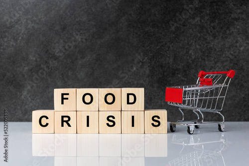 Wooden block with the word food Crisis and an empty shopping cart on a dark grey background. The concept of a global food shortage crisis with the effects of war.