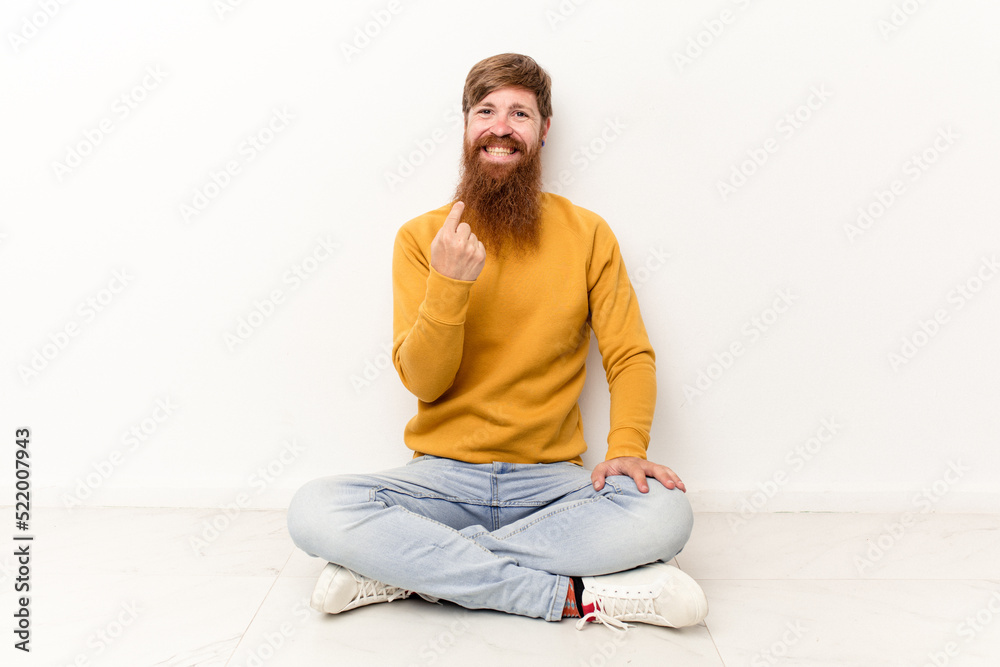 Young caucasian man sitting on the floor isolated on white background pointing with finger at you as if inviting come closer. Young caucasian man sitting on the floor isolated on white background pointing with finger at you as if inviting come closer.