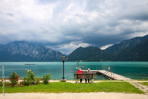 Attersee - famous Austrian turquoise lake. The view on two old ladies sitting on the wooden bench in front of the turquoise lake. Mountains in the background. Cloudy sky.