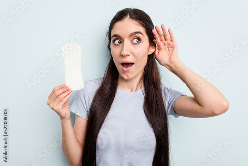 Young caucasian woman holding a sanitary napkin isolated on blue background trying to listening a gossip.