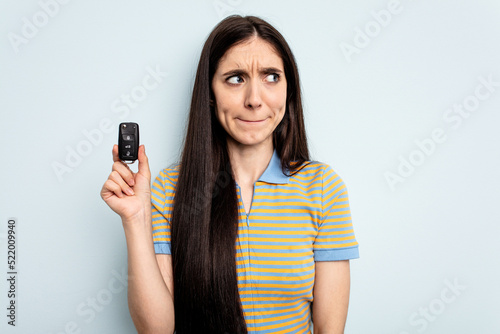 Young caucasian woman holding car keys isolated on blue background confused, feels doubtful and unsure.