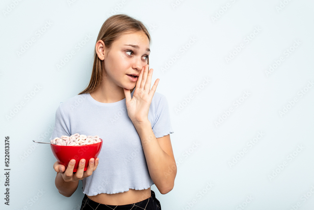 Young caucasian girl eating cereals isolated on blue background is saying a secret hot braking news and looking aside Young caucasian girl eating cereals isolated on blue background is saying a secret hot braking news and looking aside