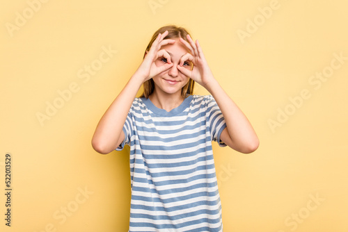 Young caucasian girl isolated on yellow background showing okay sign over eyes © Asier
