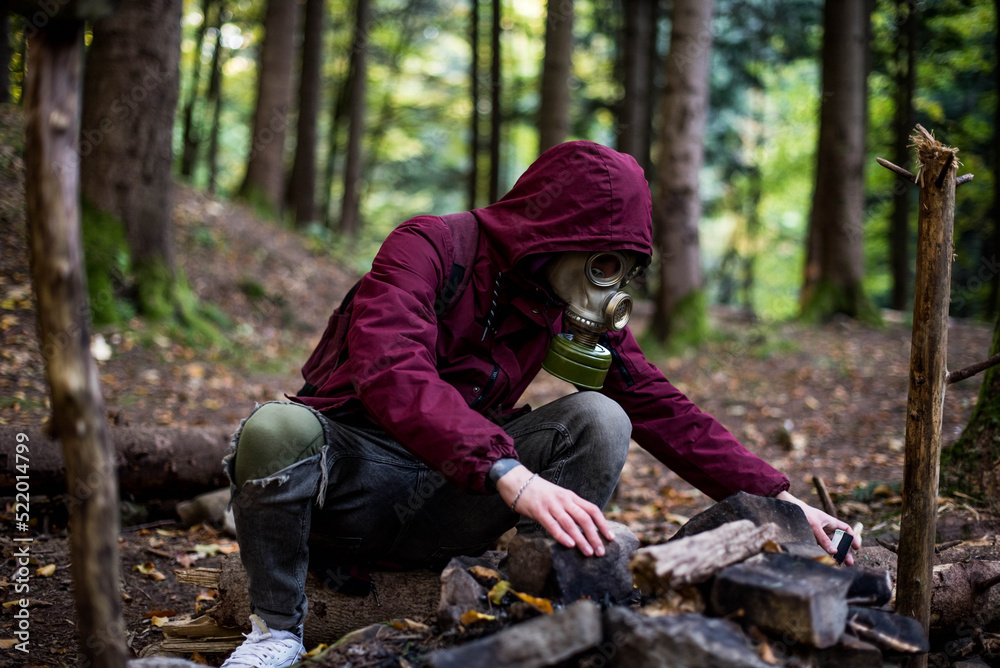 a male in a gas mask and cherry jacket sits in the middle of the forest. nuclear war concept