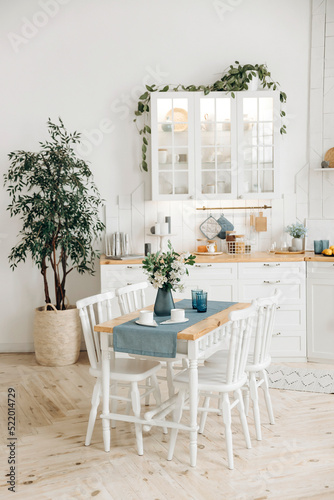 Wooden dining table with a bouquet of fresh flowers in a vase and white chairs in a modern white Scandinavian style kitchen with kitchen accessories and plants in pots. Empty space.