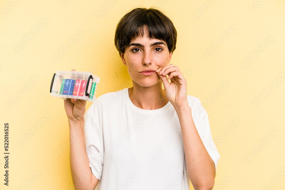 Young caucasian woman holding a batteries box isolated on yellow background with fingers on lips keeping a secret. Young caucasian woman holding a batteries box isolated on yellow background with fingers on lips keeping a secret.