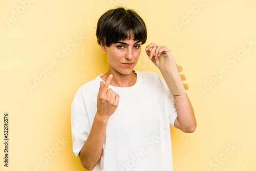 Young caucasian woman hand sling isolated on yellow background pointing with finger at you as if inviting come closer.