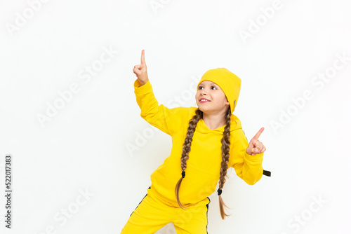 A little girl in a hat and a yellow suit points to your advertisement on a white isolated background. Autumn clothes for children. Copy space.