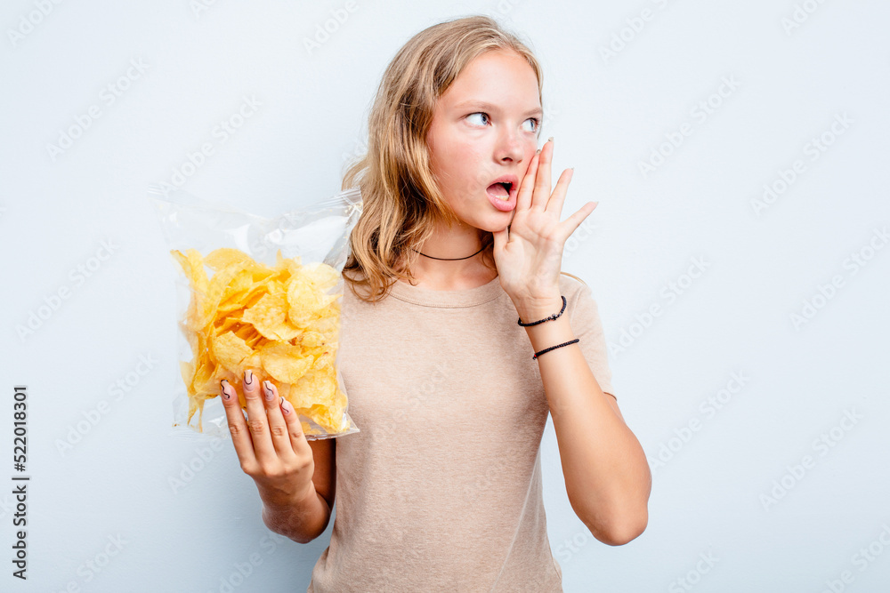 Caucasian teen girl holding bag of chips isolated on blue background shouting and holding palm near opened mouth. Caucasian teen girl holding bag of chips isolated on blue background shouting and holding palm near opened mouth.