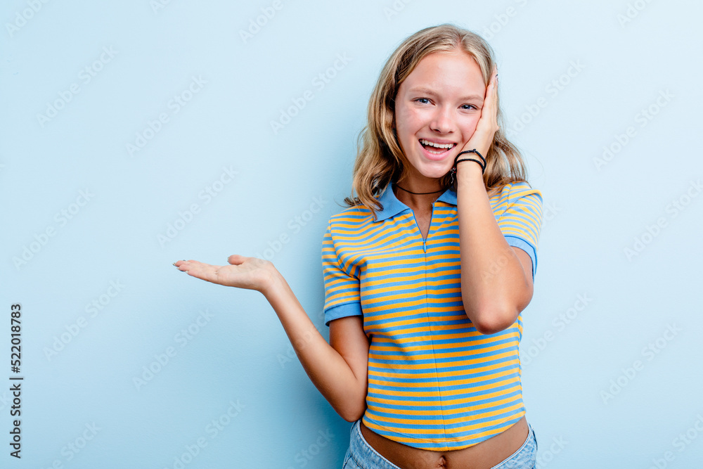 Caucasian teen girl isolated on blue background holds copy space on a palm, keep hand over cheek. Amazed and delighted. Caucasian teen girl isolated on blue background holds copy space on a palm, keep hand over cheek. Amazed and delighted.