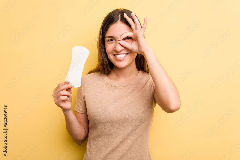 Young caucasian woman holding a sanitary napkin isolated on yellow background excited keeping ok gesture on eye. Young caucasian woman holding a sanitary napkin isolated on yellow background excited keeping ok gesture on eye.