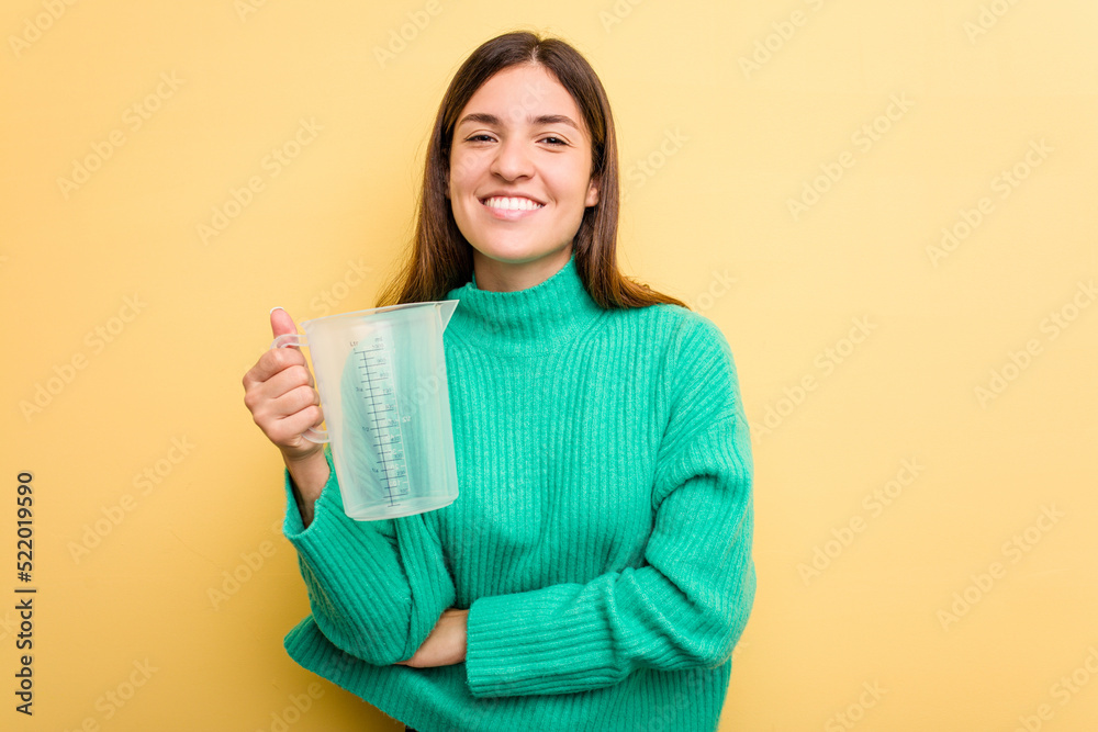 Young caucasian woman holding measuring jug isolated on yellow background laughing and having fun. Young caucasian woman holding measuring jug isolated on yellow background laughing and having fun.